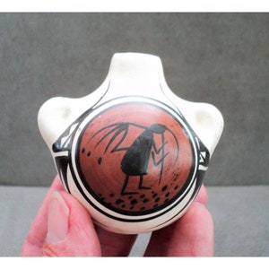 May include: A small, white ceramic pot with a red and black design. The pot has two small handles on the sides and a round, red circle in the centre with a black figure inside. The figure is a stylised human with a long, flowing robe and a staff in its hand.