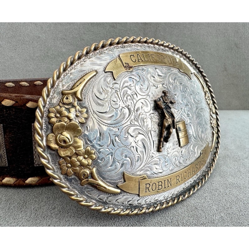 Puede incluir: Hebilla de cintur&oacute;n ovalada plateada con detalles dorados. Presenta un dise&ntilde;o floral, un jinete a caballo y las palabras "California" y "Robin Richards". La hebilla tiene un borde en forma de cuerda y est&aacute; sujeta a un cintur&oacute;n de cuero marr&oacute;n.