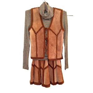 May include: A tan suede vest and matching skirt set with dark brown whipstitch detailing and lace-up front closure, layered over a long-sleeve turtleneck shirt featuring thin horizontal stripes in shades of green, red, and yellow.