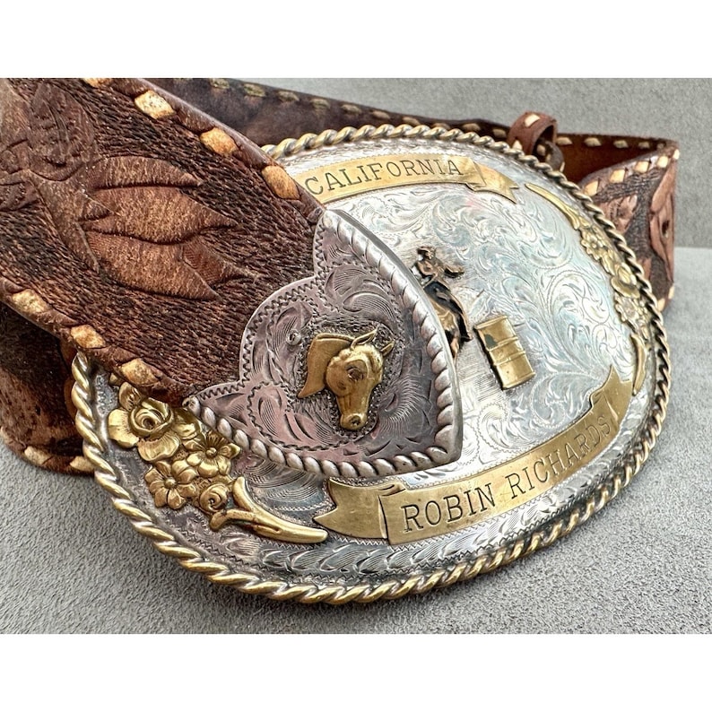 Puede incluir: Un cintur&oacute;n de cuero marr&oacute;n con una hebilla grande plateada y dorada. La hebilla presenta una cabeza de caballo, detalles florales y las palabras "California" y "Robin Richards". El cintur&oacute;n tiene un dise&ntilde;o detallado y texturizado.
