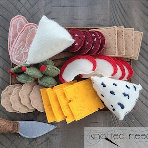 May include: A felt food play set featuring a variety of items, including slices of meat, cheese, olives, crackers, and a knife. The set includes a wooden tray and is designed for children's play. The colors are primarily pink, white, yellow, and brown.