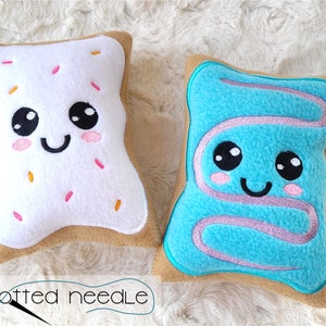 Może przedstawiać: Dwie pluszowe poduszki w kształcie Pop-Tarta, jedna z białym lukrem i posypką, a druga z niebieskim lukrem. Obie poduszki mają wyszywane oczy, uśmiech i różowe policzki.