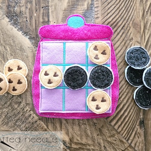 Può includere: Un gioco di barattolo di biscotti in feltro con biscotti al cioccolato e Oreo. Il tabellone di gioco è rosa con una griglia viola e i biscotti sono in feltro.