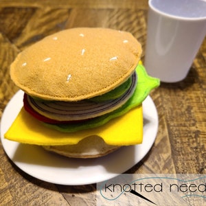 Può includere: Un hamburger di feltro con un pan al sesamo, lattuga, pomodoro, cipolla e formaggio su un piatto bianco. L'hamburger è accanto a un bicchiere d'acqua trasparente.