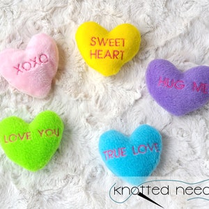 May include: Five plush heart-shaped decorations in various colors: pink with "XOXO", yellow with "SWEET HEART", purple with "HUG ME", green with "LOVE YOU", and blue with "TRUE LOVE". The hearts are on a white, textured background.