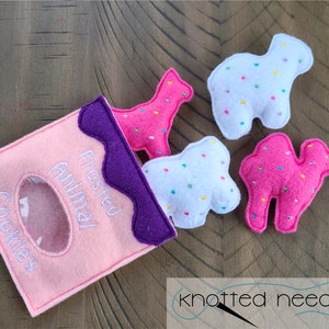 May include: A pink felt bag with the text "Frosted Animal Cookies" on the front. The bag is open and four felt animal cookies are spilling out. The cookies are pink, white, and have white sprinkles on them. The cookies are shaped like a giraffe, camel, and two different bears.