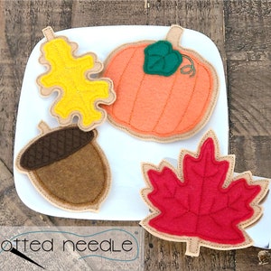 May include: A white plate displays four felt autumn-themed decorations: a yellow oak leaf, an orange pumpkin with a green stem, a brown acorn, and a red maple leaf. The decorations are outlined with a tan stitching.