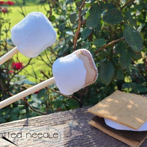 Puede incluir: Primer plano de un juego de s'mores de fieltro, que incluye dos malvaviscos blancos en brochetas de madera y un sándwich s'more. El sándwich s'more tiene una base marrón, un centro de malvavisco blanco y una parte superior de galleta graham beige. El texto "knotted needle" es visible.