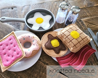 Könnte beinhalten: Filz-Spielzeug-Essen-Set mit einem rosafarbenen und weißen Donut, einem rosafarbenen und weißen Pop-Tart, einer braunen Waffel mit gelber Mitte, einem braunen Pfannkuchen mit gelber Mitte, einem roten Speckstreifen und einem weißen Ei in einer grauen Pfanne. Das Set befindet sich auf einem weißen Teller mit einem Holz-Hintergrund.