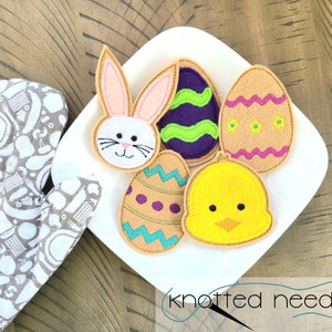 May include: Five felt Easter-themed appliques on a white plate. The appliques include a bunny, a chick, and three eggs. The eggs are decorated with different patterns, including stripes, polka dots, and swirls. The appliques are all in shades of yellow, pink, purple, green, and brown.