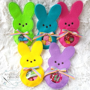 以下が含まれることがあります： 緑、青、ピンク、黄色、紫のカラフルなフェルトのウサギ型お菓子入れのコレクション。 各ウサギは、キャンディーで満たされた丸い開口部があり、虹色のリボンで飾られています。 ロゴ「knotted needle designs」は左下にあります。