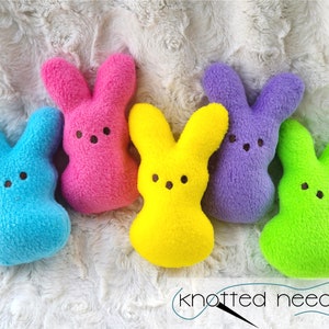 May include: Five plush bunny-shaped toys in different colors: blue, pink, yellow, purple, and green. The toys are arranged in a semi-circle on a white fluffy surface. The text "knotted needle" is visible at the bottom of the image.