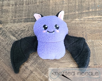 Puede incluir: Un murciélago de peluche morado con alas negras y una cara bordada linda. El murciélago tiene orejas rosas y una pequeña sonrisa.