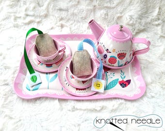 May include: A pink metal tea set with a teapot, two cups, and saucers. The set features a floral design with birds and a pink and white color scheme. The tea set is on a pink tray with a lace trim. Two tea bags are included.