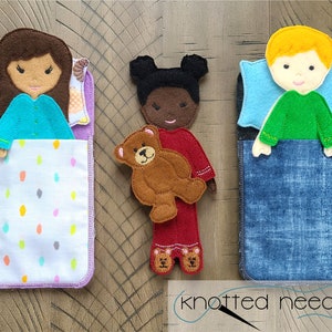 May include: Three felt dolls in bed, one with a brown teddy bear. The dolls are wearing different coloured pyjamas and have different skin tones. The beds are made of felt and have different patterns.