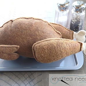 Może przedstawiać: Filcowy indyk na szarej tacy, z dwiema solniczkami i pieprzniczkami w tle. Indyk jest brązowy z białymi filcowymi pałeczkami. Taca stoi na drewnianej powierzchni.