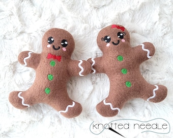 May include: Two gingerbread people plush toys, one with a red bow tie and the other with a red bow, on a white fluffy surface.