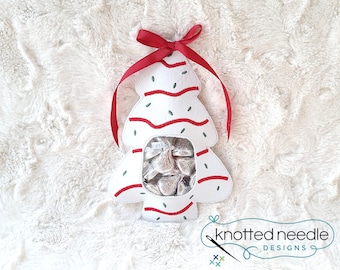 May include: A white, tree-shaped felt pouch featuring red embroidered wavy lines and small green stitched accents. The center has a clear plastic window revealing silver-wrapped chocolate candies inside. A red ribbon is tied at the top.