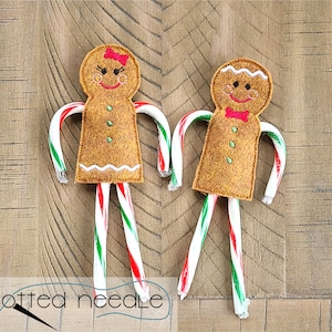 May include: Two felt gingerbread people with candy cane arms and legs. One has a red bow and eyelashes, the other a red bow tie. Both have embroidered smiles and button details. The background is a wooden surface.