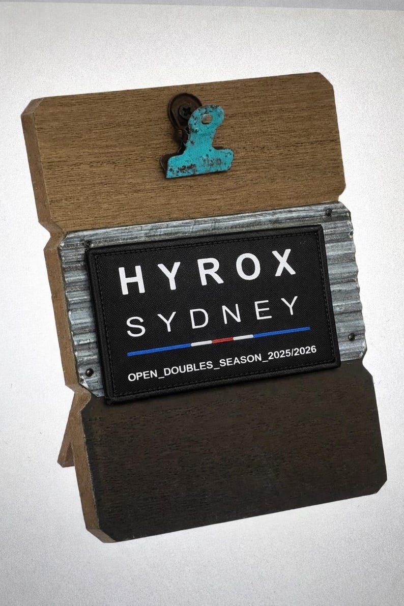 Puede incluir: Un organizador de escritorio de madera con un acabado envejecido. Presenta un parche rectangular negro con el texto "HYROX SYDNEY" en blanco y "OPEN_DOUBLES_SEASON_2025/2026". Un clip turquesa est&aacute; sujeto en la parte superior.