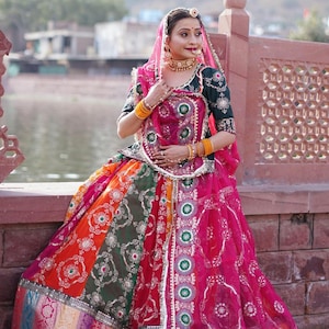 May include: A woman wearing a traditional Indian outfit with a long, colorful skirt, a green blouse, and a pink veil. She is wearing gold jewelry and has her hair pulled back.