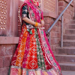 May include: A woman wearing a traditional Indian outfit with a colorful skirt, a green blouse, and a pink dupatta. She is standing in front of a brick wall and has a gold necklace and bangles on.