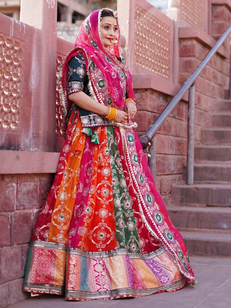 May include: A woman wearing a traditional Indian outfit with a colorful skirt, a green blouse, and a pink dupatta. She is standing in front of a brick wall and has a gold necklace and bangles on.