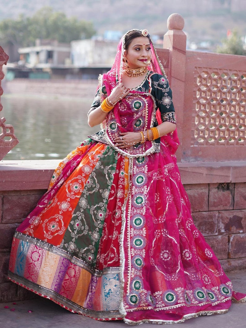 May include: A woman wearing a traditional Indian outfit with a long, colorful skirt, a green blouse, and a pink veil. She is wearing gold jewelry and has her hair pulled back.