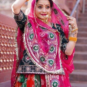 May include: A woman wearing a traditional Indian outfit with a pink dupatta, green and gold embroidered choli, and a colorful skirt. She is wearing jewelry, including a nose ring and a necklace.