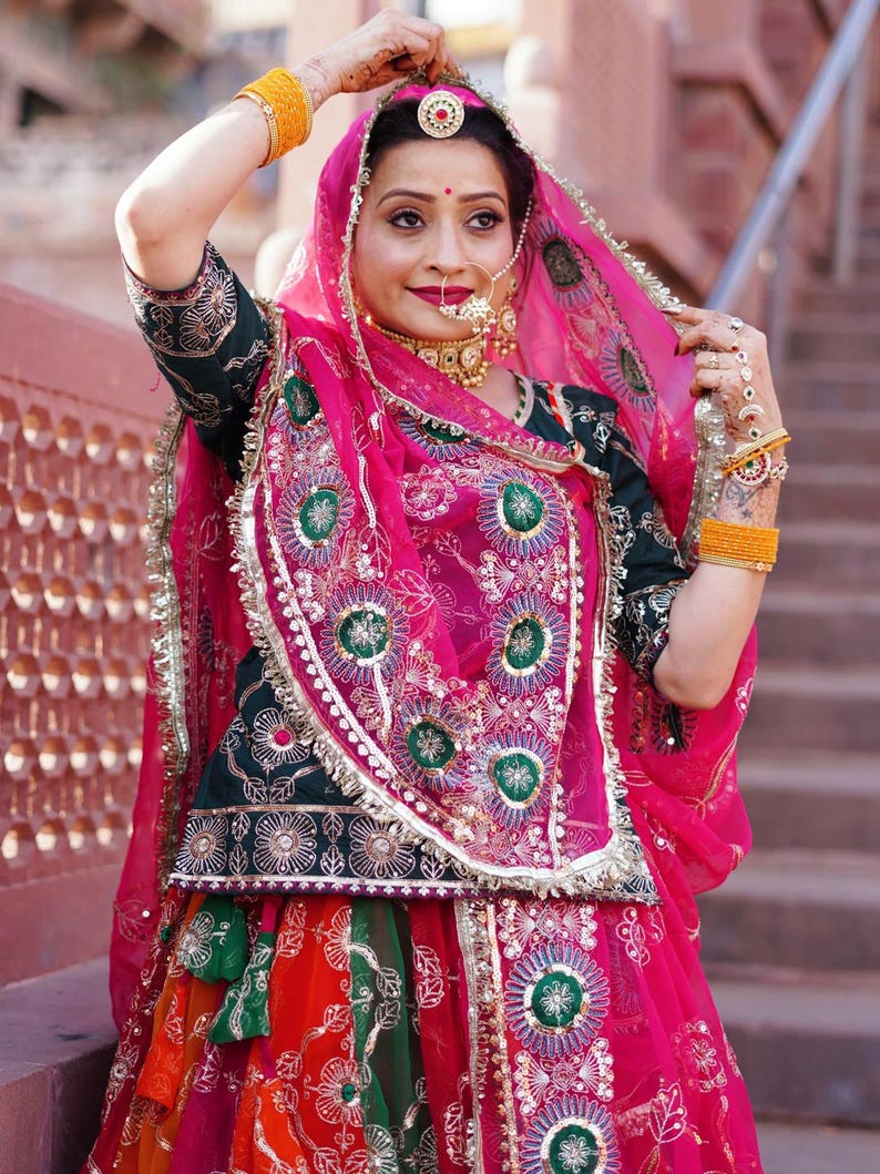 May include: A woman wearing a traditional Indian outfit with a pink dupatta, green and gold embroidered choli, and a colorful skirt. She is wearing jewelry, including a nose ring and a necklace.