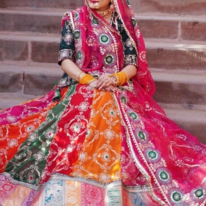 May include: A woman wearing a colorful, embroidered skirt and a pink dupatta with gold accents. She is sitting on a set of stone steps.