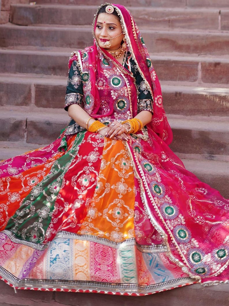 May include: A woman wearing a colorful, embroidered skirt and a pink dupatta with gold accents. She is sitting on a set of stone steps.