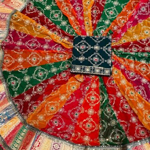 May include: A colorful circular skirt with a green embroidered top. The skirt has a multi-colored border with gold accents. The skirt is made of a sheer fabric with a floral pattern.