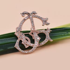 May include: A rose gold brooch in the shape of the intertwined letters "DG", encrusted with small, sparkling stones. The brooch is set against a background of green leaves and a pale pink surface. The brand name "ANRII JEWELS" is visible.