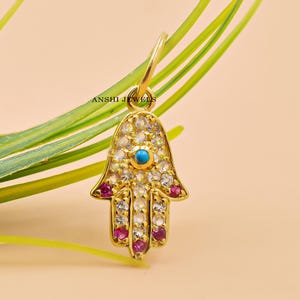 Op de afbeelding: Gouden hamsa handbedel met een turquoise steen in het midden en roze stenen aan de randen. De bedel is bedekt met kleine witte stenen.