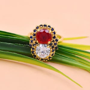 K&ouml;nnte beinhalten: Ein Goldring mit einer vertikalen Anordnung aus zwei gro&szlig;en runden Steinen, einem tiefroten und einem klaren wei&szlig;en, umgeben von einem Kranz aus kleinen dunkelblauen Steinen. Der Ring sitzt auf einer d&uuml;nnen Goldschiene mit polierter Oberfl&auml;che.