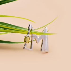 May include: Silver and gold letter M charm pendant. The pendant is in the shape of the letter M and has a silver finish. The pendant is attached to a gold chain.