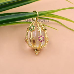 May include: Gold-colored pendant with a clear, oval-shaped center. The pendant is adorned with small, colorful gemstones and floral designs. The text "ANS JEWELS" is visible on the pendant's loop. The pendant is hanging on a green leaf.