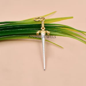 Pode incluir: Um pingente de espada dourada e prateada com uma lâmina texturizada e uma guarda dourada. O pingente é preso a um pequeno anel dourado. A espada é exibida em um leito de folhas verdes sobre um fundo marrom claro. O texto "ANSHI JEWELS" é visível.