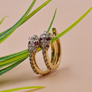 May include: A pair of gold hoop earrings with a red and white stone design. The earrings are set against a light pink background.