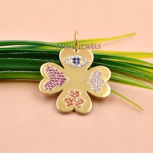 May include: A gold-toned four-leaf clover pendant featuring inlaid gemstones. The design includes a blue and white evil eye, a pink heart, a white hamsa hand, and an orange shamrock. The text ANSHI JEWELS is printed on the metal surface.