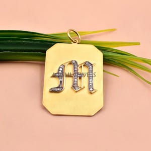 May include: A rectangular gold pendant featuring a gothic-style letter M encrusted with small clear stones. The pendant has a smooth matte finish and a small circular bail at the top for attachment. The text AISHI JEWELS is visible on the letter.