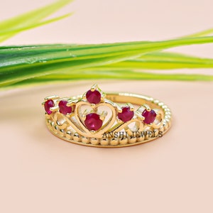 Könnte beinhalten: Goldfarbener Ring mit einem Kronendesign mit fünf roten Edelsteinen. Der Ring hat ein Perlenband und der Text "ANSHI JEWELS" ist auf dem Band sichtbar.
