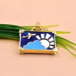 May include: A rectangular gold-tone pendant featuring an enamel design of a blue sky with a white sun, a light blue cloud, and a small airplane. The pendant has a gold bail at the top and the text ANSHJEWELS is visible on the metal surface.