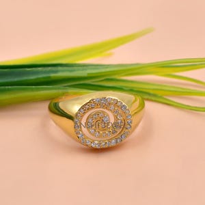 Puede incluir: Un anillo de tono dorado que presenta una cara circular con un patr&oacute;n en espiral incrustado con peque&ntilde;as piedras claras y brillantes. La banda es lisa y pulida, mostrando un acabado met&aacute;lico continuo que complementa el dise&ntilde;o central.