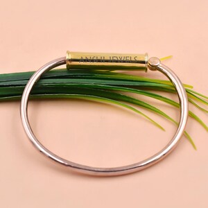 May include: A circular bangle bracelet featuring a polished rose gold-toned band and a central gold-toned cylindrical bar. The bar is engraved with the text ANSHI JEWELS. The bracelet has a smooth, metallic finish and a simple, continuous design.