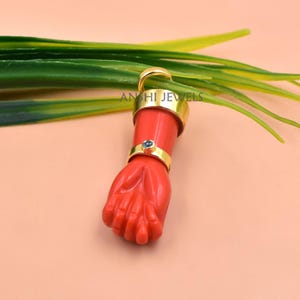 Puede incluir: Un colgante con forma de puño cerrado tallado en material rojo, con un brazalete de metal dorado en la muñeca y una pequeña piedra azul incrustada. La pieza lleva la inscripción ANSHI JEWELS e incluye una anilla de metal dorado.