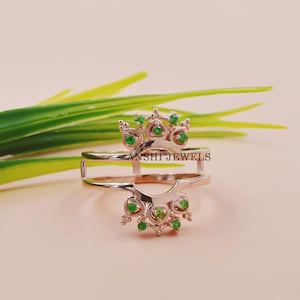 Puede incluir: Un anillo de oro rosa con un diseño de doble banda. La banda superior presenta un diseño delicado con piedras preciosas verdes. El texto "ANSHI JEWELS" es visible en el anillo.