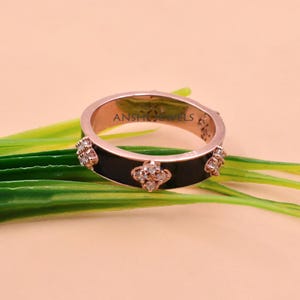 May include: A rose gold band ring featuring a black enamel inlay and repeating four-petaled flower motifs set with small clear stones. The interior of the band is engraved with the text ANSH JEWELS. The ring has a polished metallic finish.