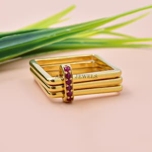 May include: A gold-colored ring set with a row of red gemstones. The ring has a square design and is stacked with three additional rings. The text "AN JEWELS" is visible on the ring. The background is a light pink color.